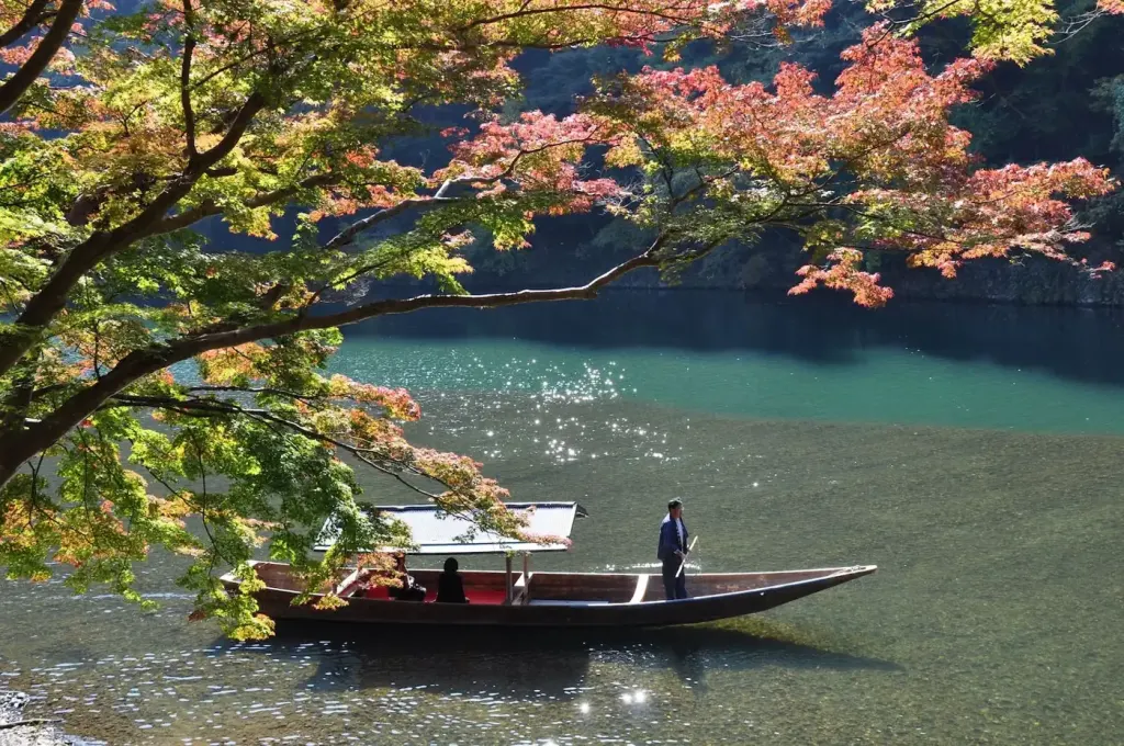 A boatman poling a wooden boat beneath autumn maples on the river at Arashiyama